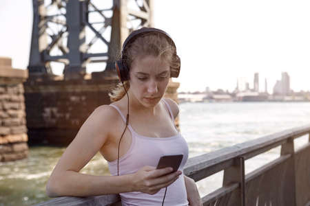 Young woman beside river, wearing headphones, using smartphone, New York City, USAの写真素材