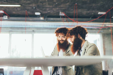Male hipster twins working on laptop at office deskの写真素材