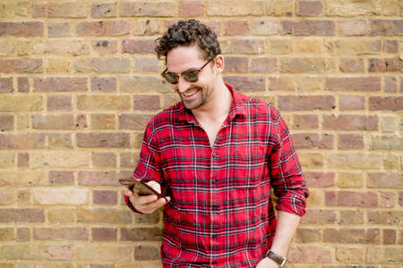 Young man leaning against brick wall reading smartphone texts