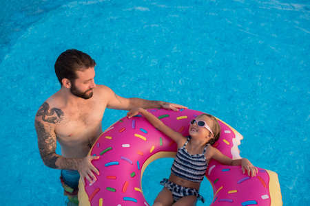 Young girl in swimming pool, relaxing on inflatable ring, father standing beside herの写真素材