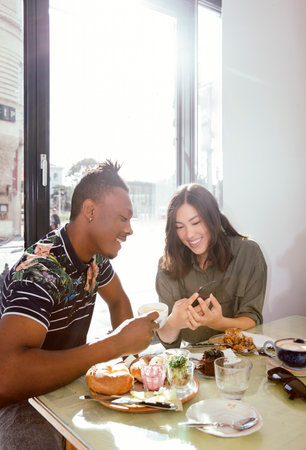 Smiling couple enjoying breakfast and looking at smart phone in restaurantの写真素材