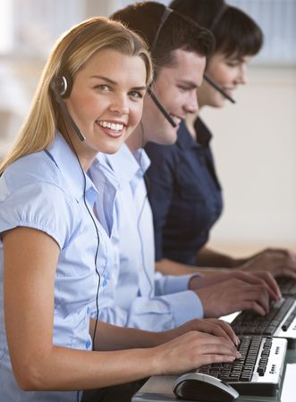A group of people are typing on keyboards and wearing headsets.  The young woman is smiling at the camera.  Vertically framed shot.の写真素材