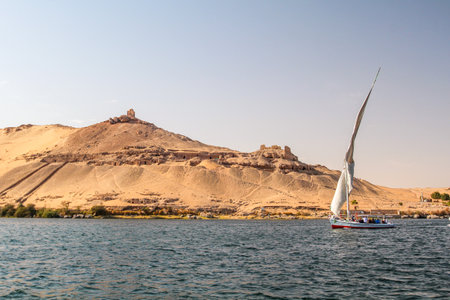 Sailboat on the Nile river at the shore of Aswan, tombs of the nobles in the background, Egyptの写真素材