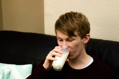 A handsome young blonde man drinking milk while sat on the sofaの写真素材