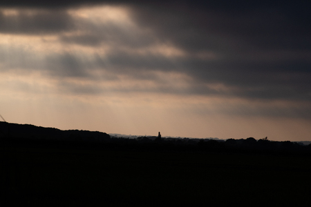 silhouettes of bushes and trees on the horizon with sunbeams shining through the coulds at sunsetの写真素材