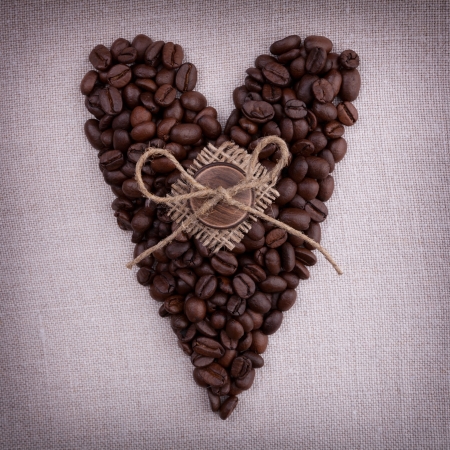 Dark roasted coffee beans  in the shape of a heart with wooden button and bow  on it on fabric backgroundの写真素材