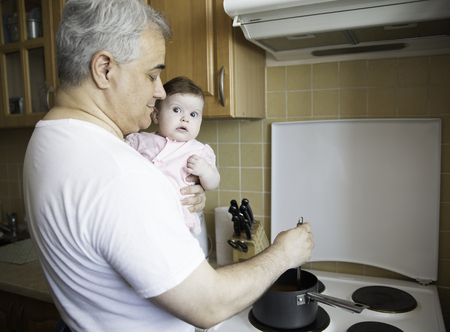 Man in the kitchen holding a child on his shoulder, cooking in the soup pot. 6-months old adorable baby, bonding concept photo, familyの写真素材