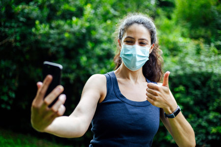 A young caucasian woman wearing a mask with phone taking selfie in the park