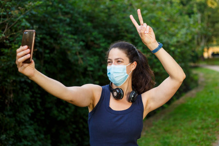 A young caucasian woman wearing a mask with headphones and smart phone taking selfie in the park
