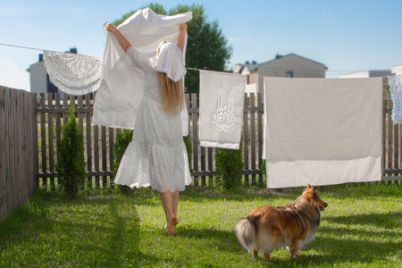 Beautiful young blonde woman back view hanging and drying her clean white laundry on rope with collie dog sitting aroundの写真素材