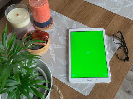 White tablet with green screen laying on wooden table next to candle, natural stones and green plant. Home office, interior with working table.の写真素材