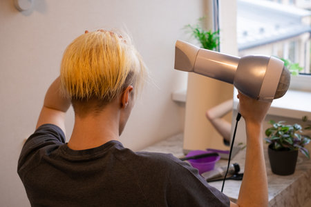 back view. Young guy sitting in front of the mirror and drying his hair by himself at home. beauty salon at home concept. The process of hair coloring, Hair Coloring in process, Man gets new hair colour.の写真素材