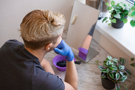 back view. Young guy in blue gloves sitting in front of the mirror and coloring his hair by himself at home. Beauty salon at home concept. The process of hair coloring, Hair Coloring in process, Man gets new hair colour.の写真素材