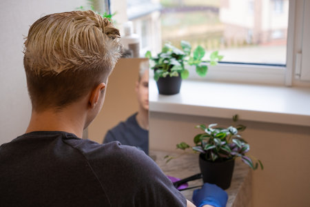 back view. Young guy in blue gloves sitting in front of the mirror and coloring his hair by himself at home. Beauty salon at home concept. The process of hair coloring, Hair Coloring in process, Man gets new hair colour.の写真素材