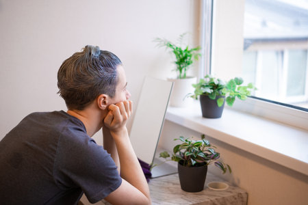 back view. Young guy with applied hair dye sitting in front of mirror and looking at window thinking. beauty salon at home concept. The process of hair coloring, Hair Coloring in process, Man gets new hair colour.の写真素材