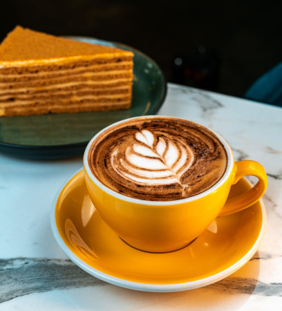 Hot Mocha with Leaf Latte Art in Yellow Cup on Marble Table. Blurred slice of layered dessert in...