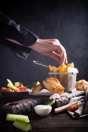Fish and chips. Fried fish fillet with homemade baked potatoes and fresh salad on wooden cutting board with human hand.の写真素材
