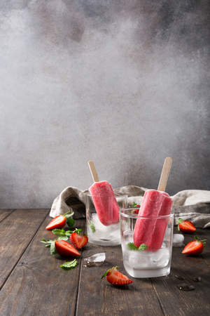 Homemade strawberry popsicles in glass with ice, strawberries and mint on old wooden background. Healthy summer food concept with copy space.の写真素材