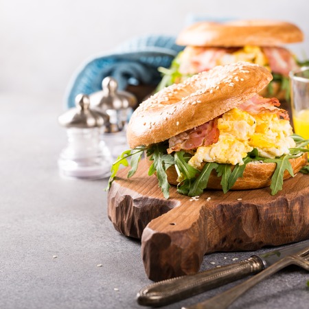 Healthy freshly baked bagel filled with scrambled eggs, rucola and fried bacon. Served on small wooden cutting board on gray background. Breakfast food. Copy space.の写真素材
