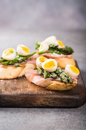 Easter breakfast or lunch with sendwich with ham, asparagus and quail eggs on old wooden chopping board. Spring food concept with copy space.の写真素材