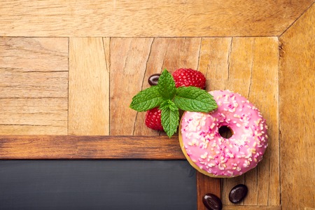 Black chalkboard with pink glazed donut, raspberries and mint on old wooden table. Food background concept for menu, cafe, restaurant. Copy space, flat lay, top view.の写真素材