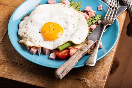 Healthy breakfast or lunch with fried egg, bread toast, green asparagus and tomatoes on blue plate on old wooden tableの写真素材