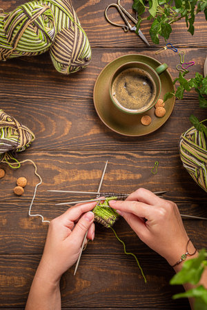 Female hands knitting green socks in progress. Flat lay on wooden table with coffee and house...