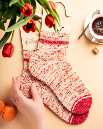 Handknitted spring wool socks with red accents on wooden sock blockers, surrounded by tulips, coffee, and macarons, creating a cozy and aesthetic composition. With woman handの写真素材