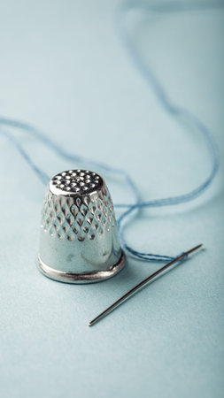 Close-up of a silver thimble and needle with blue thread resting on a soft pastel background, ideal...