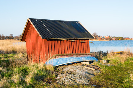 Old red boathouse by the sea  Boat in front on land  Granite rock and grass  Early morning sunshine from left の写真素材