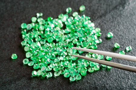 Pile of green, round cut emeralds on black stone plate. One emerald held by tweezers.の写真素材