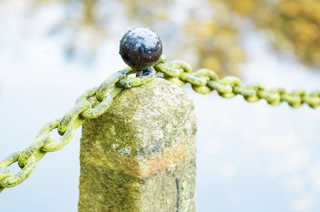 Black round knob on granite stone pillar with old moss covered iron chain. Water reflection out of focus in background. Reflections of sky in knob.の写真素材