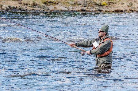 MORRUM, SWEDEN - MARCH 28, 2015: Unknown person fly fishing for trout or salmon on premiere day. Person gripping rod with both hands. River bank in background. Hip deep in water.のeditorial素材