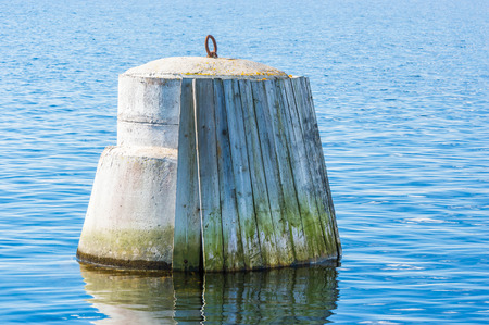 Concrete ship guard with wooden planks to protect the incoming ships from being damaged. Iron ring to moor the ship with on top. Planks are green with algae and weathered. Surrounded by sea water.の写真素材
