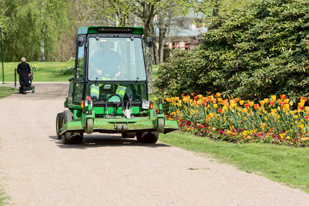 Ronneby, Sweden - May 11, 2015: John Deere grass cutting vehicle driving along gravel road in public park. Tulips and Rhododendron grow beside road.のeditorial素材