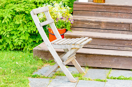 Old wooden chair in front og wooden staicase. Grass and shrubs in background. Red plastic bucket with flowers on stairs.の写真素材