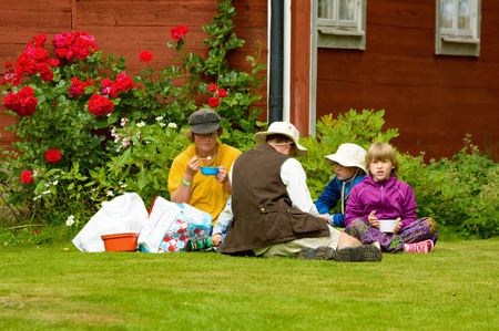 Smaland, Sweden - July 24, 2015: Family having a picnic in the lawn outside a red wooden building with some red roses. One person look at you. They sit directly on the grass without a blanket.のeditorial素材
