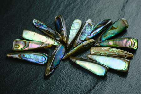 Some elongated drop shaped abalone shell beads that show lots of iridescent shimmer in different colors. Photographed on black stone surface. Abalone is a source for mother of pearl.の写真素材