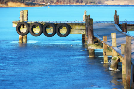 A small wooden pier stretching out into the icy sea. The blue sky is reflected in the ice. Rubber...