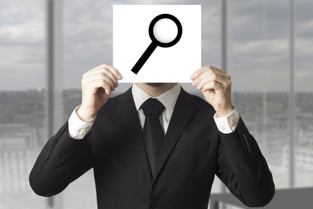 businessman in black suit  hiding face behind sign loup magnifierの写真素材