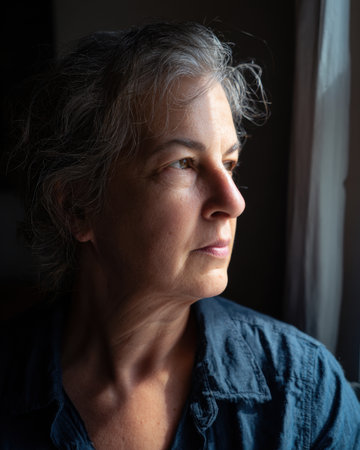 A thoughtful middle aged woman gazes out of window, capturing moment of reflection and serenityの素材