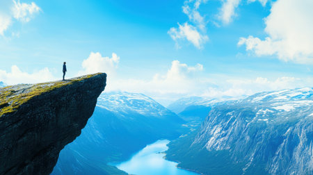 A solo traveler stands at edge of cliff, overlooking breathtaking fjord surrounded by majestic mountains and clear blue skiesの素材