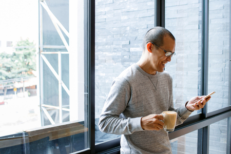 Cheerful asian male small business owner with coffee cup in hands searching on phone, reading incoming sms message on smartphone while standing in his office.  Copy space.の写真素材