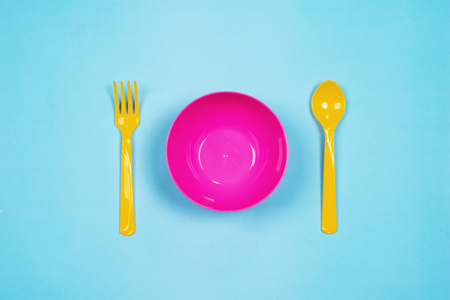 Set of empty pink plastic tableware bowls, yellow spoons and forks  isolated on blue background. Flat lay, Top view. Minimal style.の写真素材
