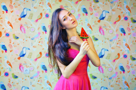 Pretty brunette european woman with long wavy hair, holding in hand sweet candy, big watermelon on stick. Wearing short pink dress. Posing at studioの写真素材