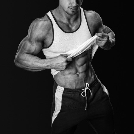 Athletic male model posing in sports clothing, pulling up tank top to reveal fit muscular abs. Isolated on black backgroundの写真素材