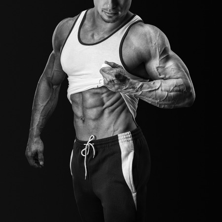Handsome bodybuilder shows his great physique, perfect shoulders, biceps, triceps and pulling up tank top to reveal fit muscular abs. Isolated on black backgroundの写真素材