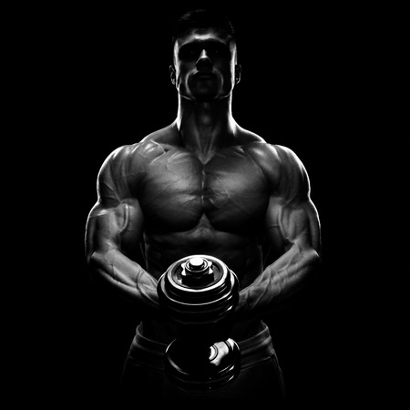 Silhouette of a bodybuilder. Power athletic man pumping up muscles with dumbbell. Confident young fitness man with strong core muscles, power hands and clenched fists. Dramatic light.の写真素材