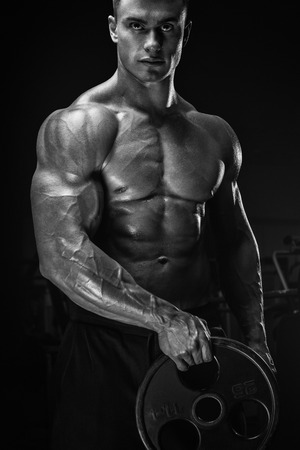 Confident handsome young athletic man doing exercises with barbell plate. Muscular body on dark background. Fitness sports. Healthcare, bodycare and physique concept.の写真素材