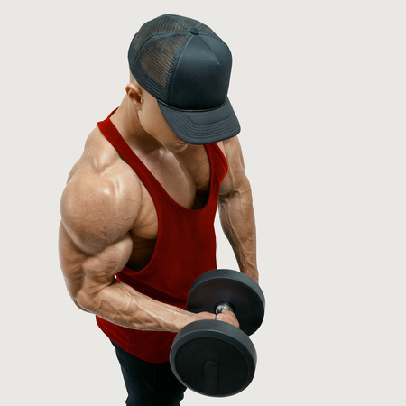 Close up shot of muscular bodybuilder wearing a red tank top and black cap doing biceps curl with dumbbell against white background. Image with clipping pathの写真素材